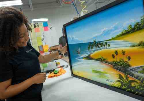 A team member frames a colorful canvas in a black wooden frame. A team member frames a colorful canvas in a black wooden frame.