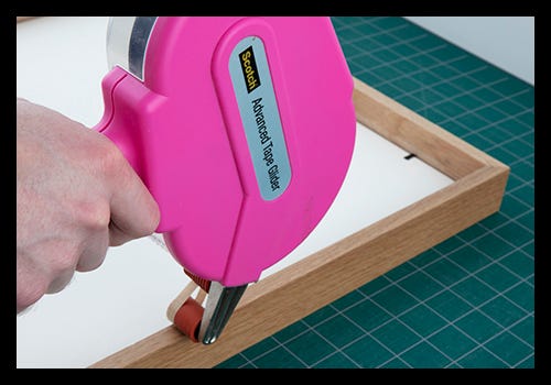 Using an adhesive tape applicator to apply tape to the back of a wooden frame Using an adhesive tape applicator to apply tape to the back of a wooden frame