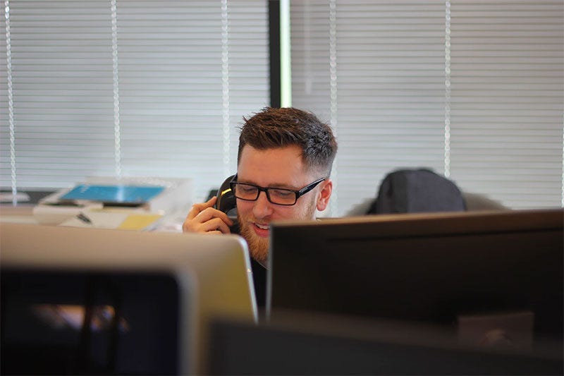 Man with glasses answering a phone in an office sitting in front of two computer monitors